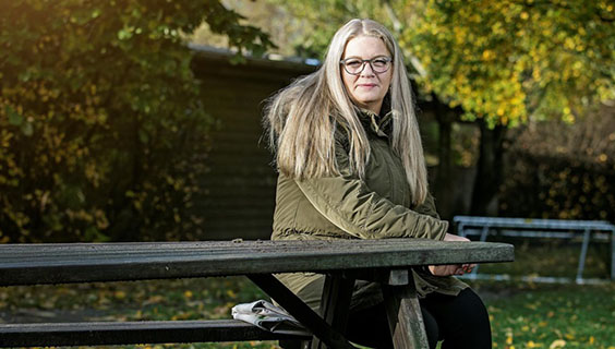 Kvinde med langt blond hår og briller sidder udendørs ved et bord med efterårstræer i baggrunden